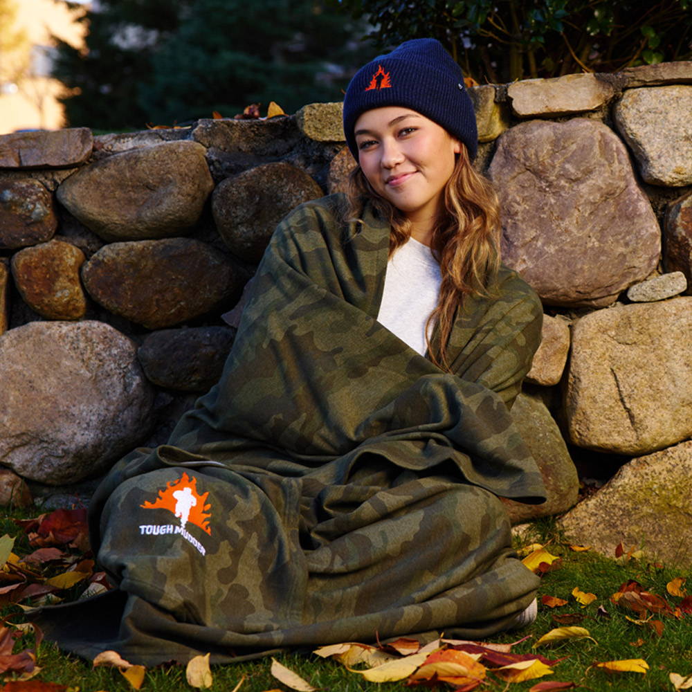 An image of a female model wearing a navy blue Tough Mudder beanie, sitting in front of a rock wall wrapped in a Tough Mudder fleece camo blanket.