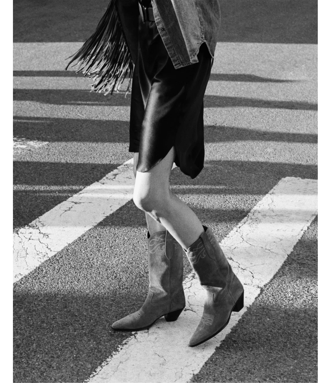 Woman walking in a crosswalk wearing a black dress and cowboy boots.