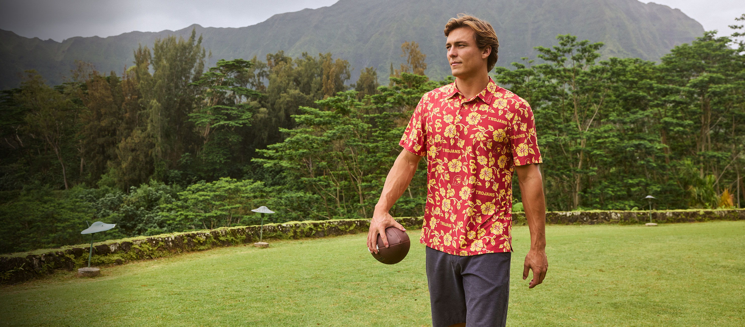 Man in a floral shirt holding a ball in a lush outdoor setting.