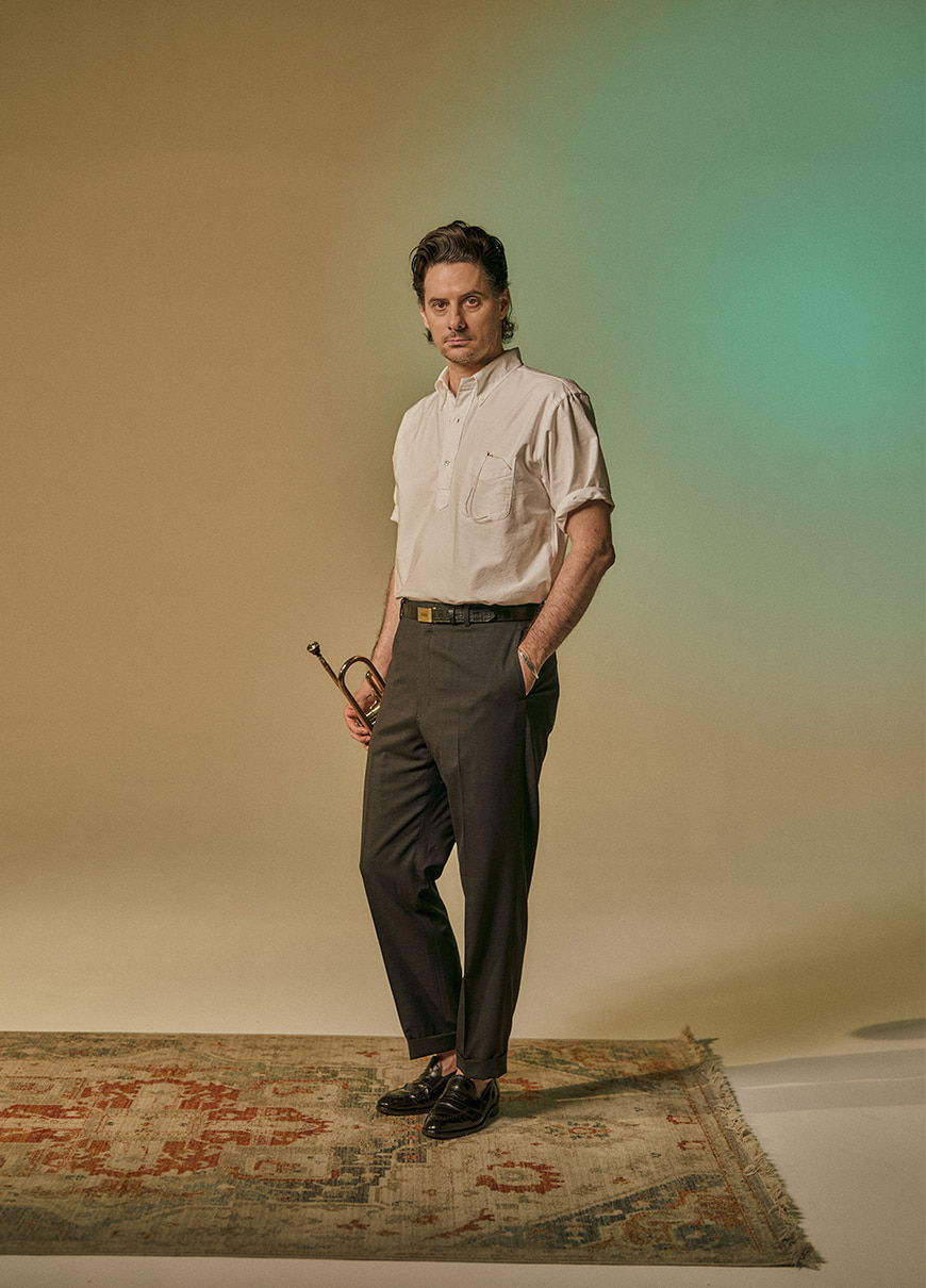 Man in a white shirt holding a trumpet, standing on a patterned rug.