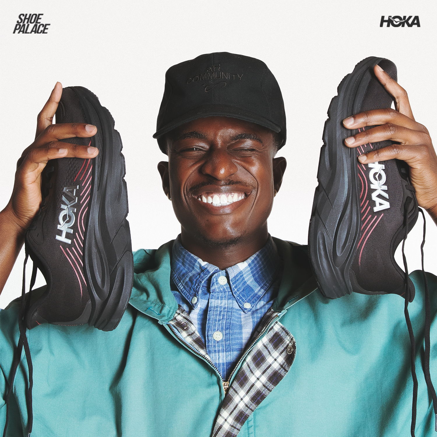 Person smiling while holding HOKA shoes, wearing a green jacket and cap.