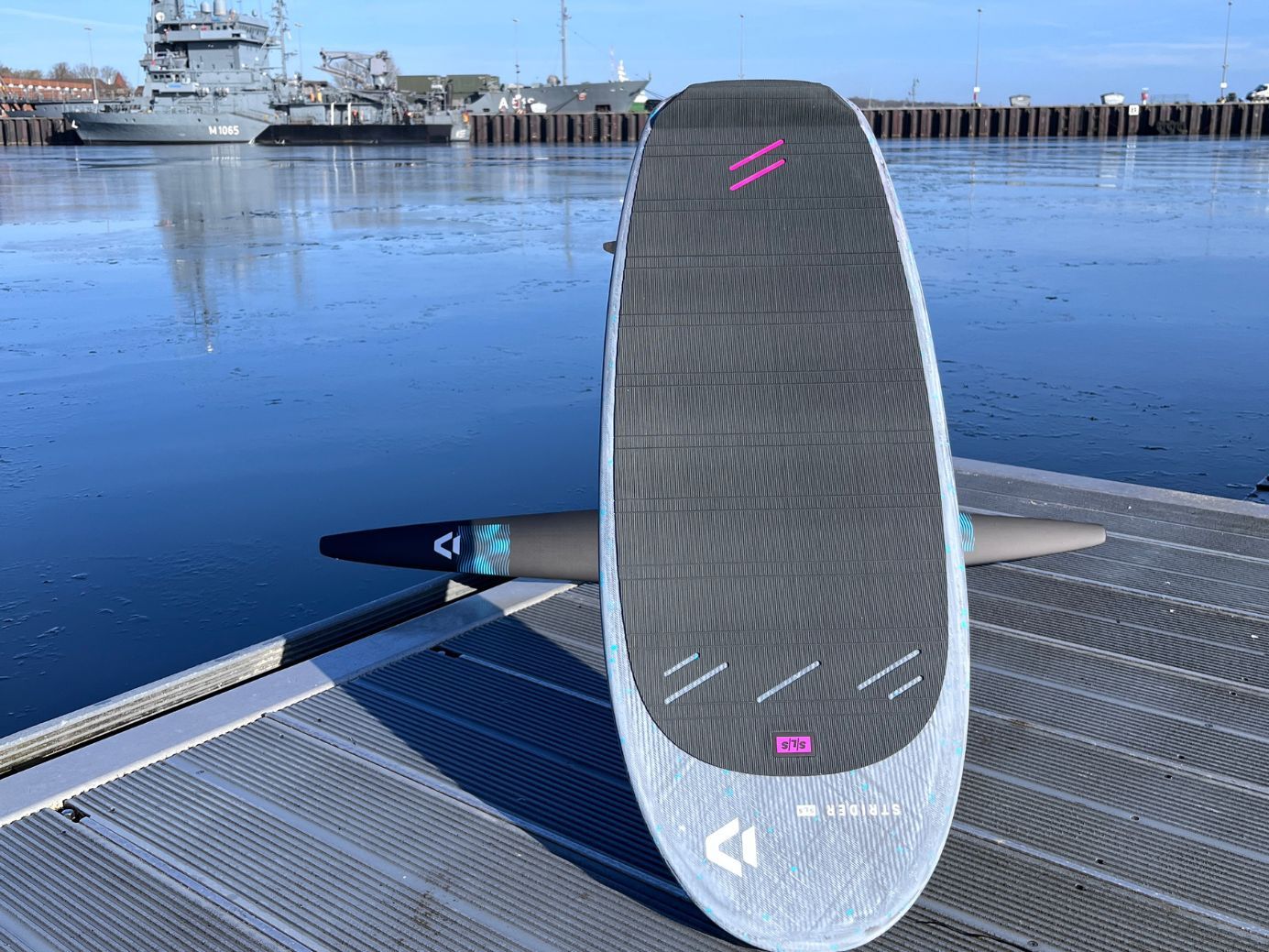 A surfboard on a dock with water and boats in the background.