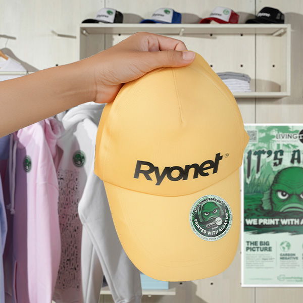 Yellow cap with 'Ryonet' branding & Living Ink Sticker held by a hand against a background of clothes and a Living Ink It's Alive poster.