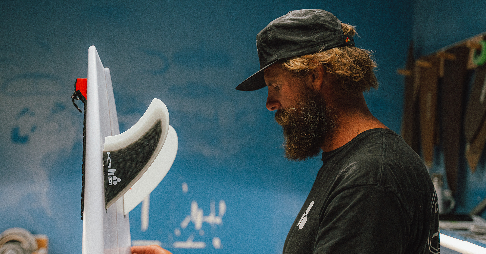 Person working on a surfboard in a workshop with blue walls.