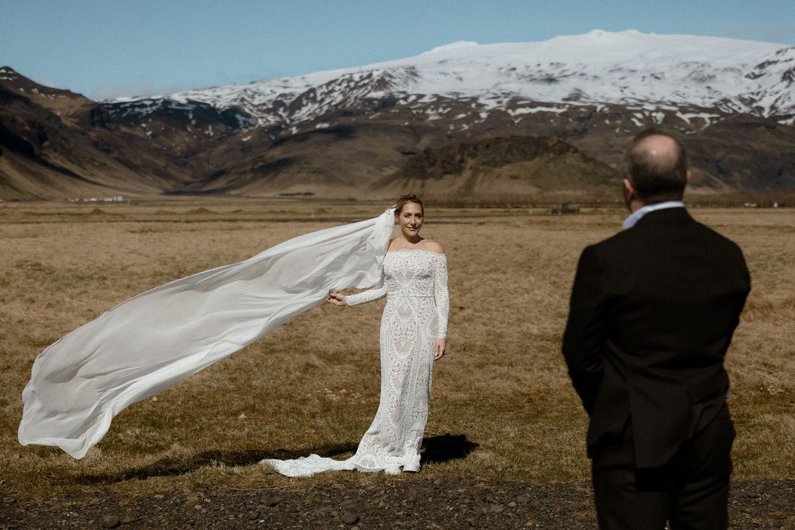 Bride in white dress with flowing veil, standing in a scenic landscape.