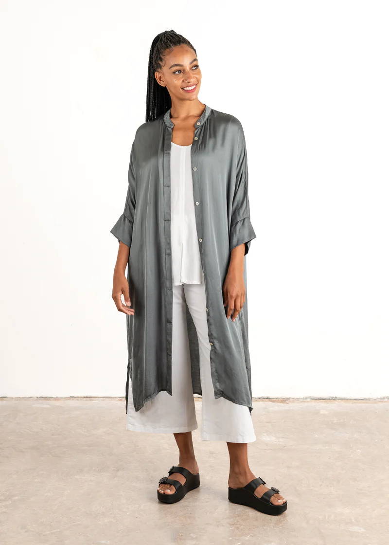 A model wearing a blue grey long satin shirt over a white top and white wide leg trousers and black chunky slides