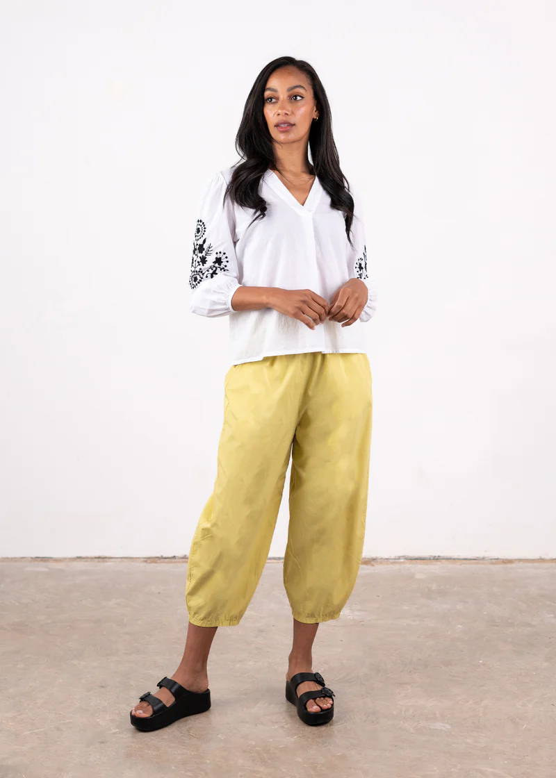 A model wearing a white long sleeved blouse with black embroidered detailing on the outer arm, with pale yellow barrel leg trousers and black chunky slides