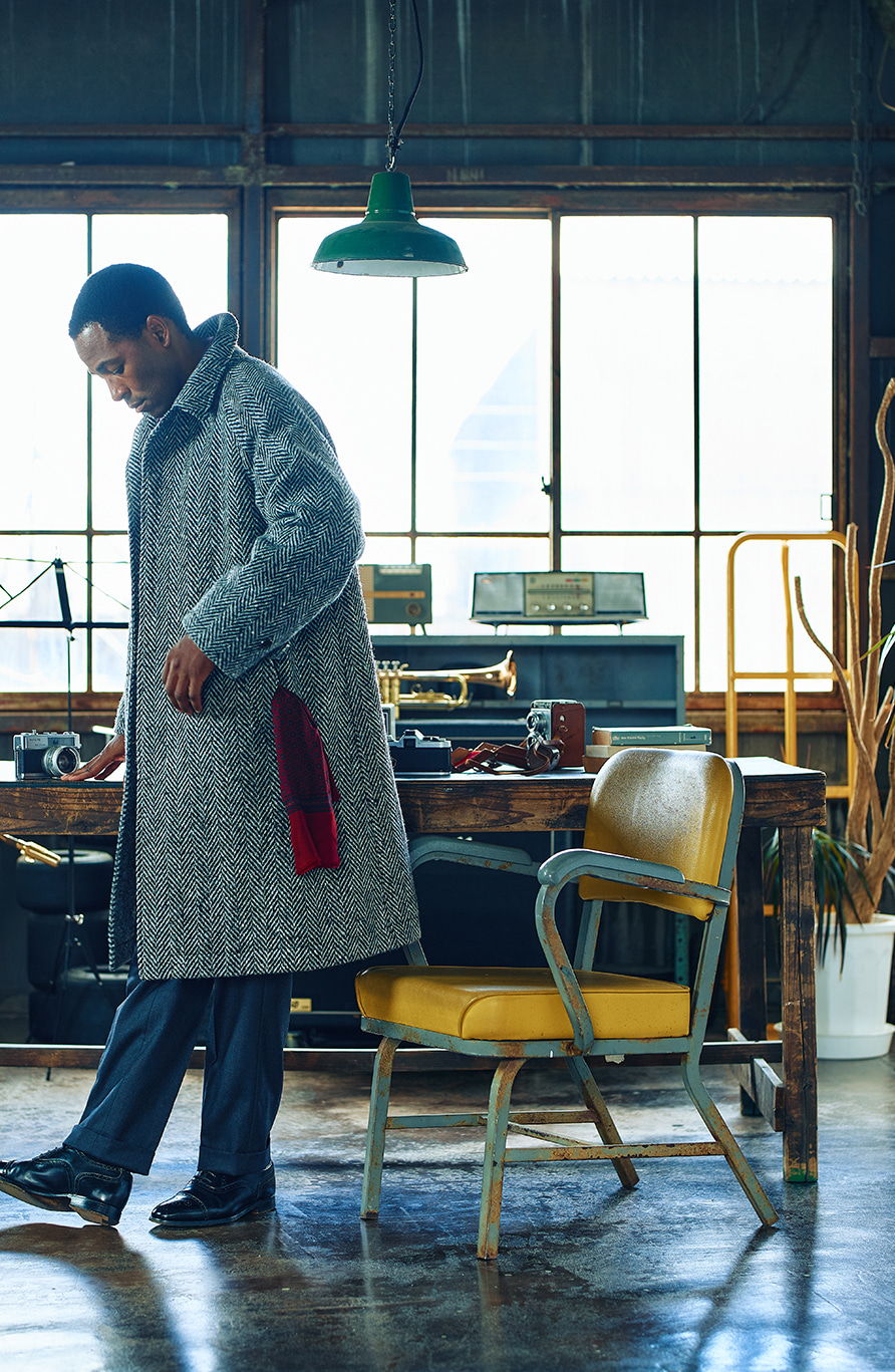 Man in a gray coat standing beside a yellow chair in a workshop.