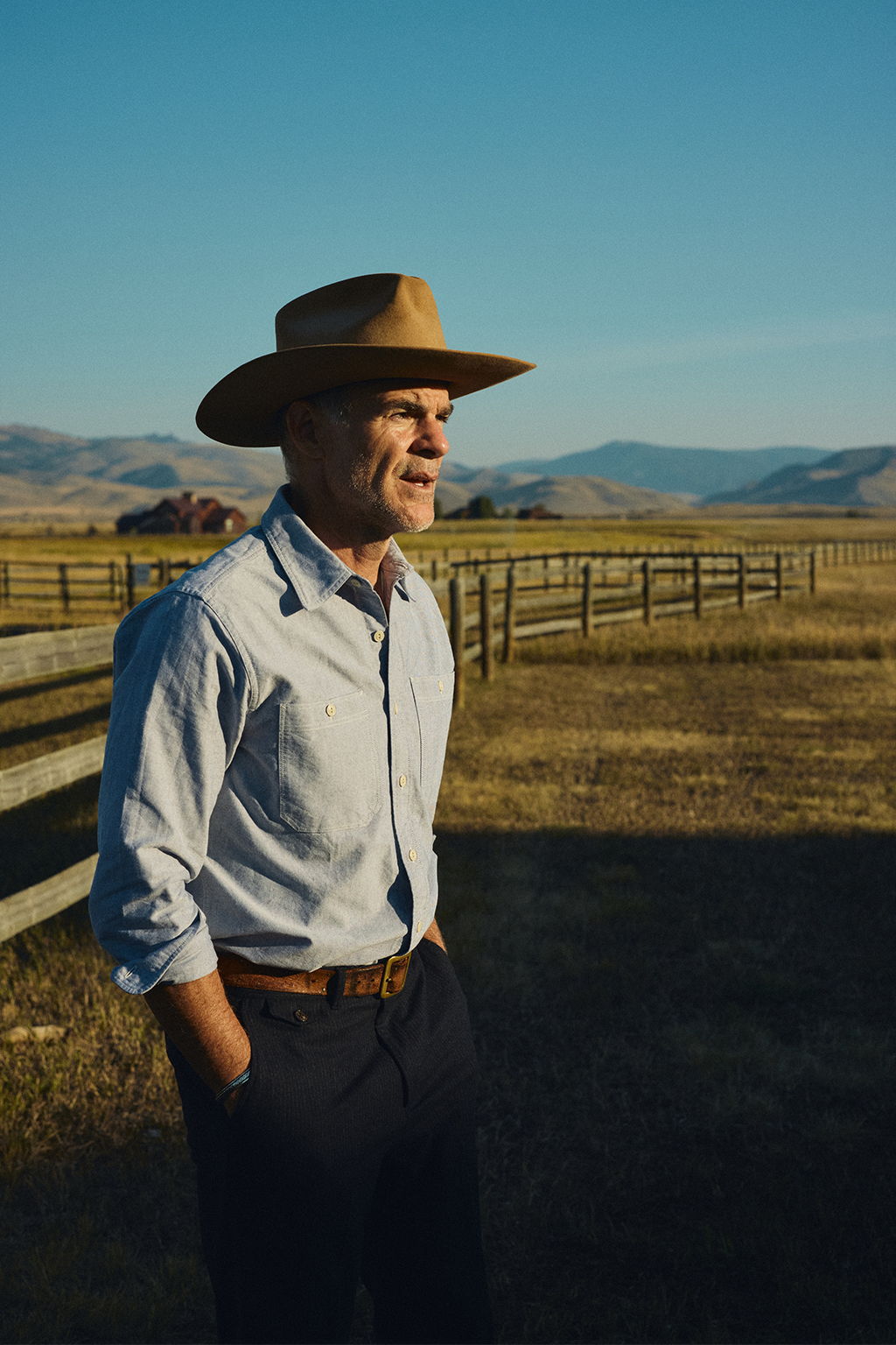 Michael Kelly standing next to a fence in Pray, Montana wearing a vintage stetson, the Rivay Bridges Oxford Workshirt in Navy, Rivay Italian Wool Suit in Navy Chalk Stripe Trousers