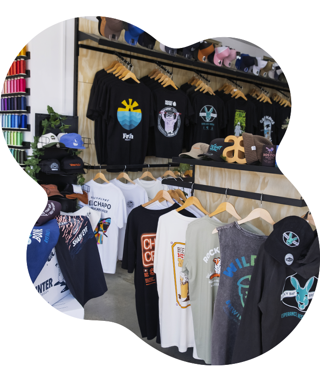 Interior of The Fabric Printer showroom displaying black t-shirts with colourful printed graphics hanging on a wall-mounted rack, alongside caps and accessories on shelving, with a rail of white and printed garments below