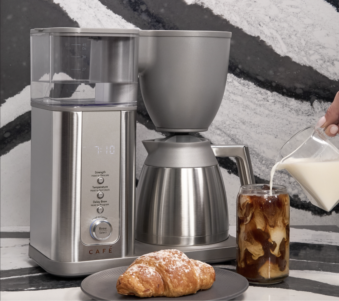 Coffee maker with croissant and iced coffee; milk being poured into glass.