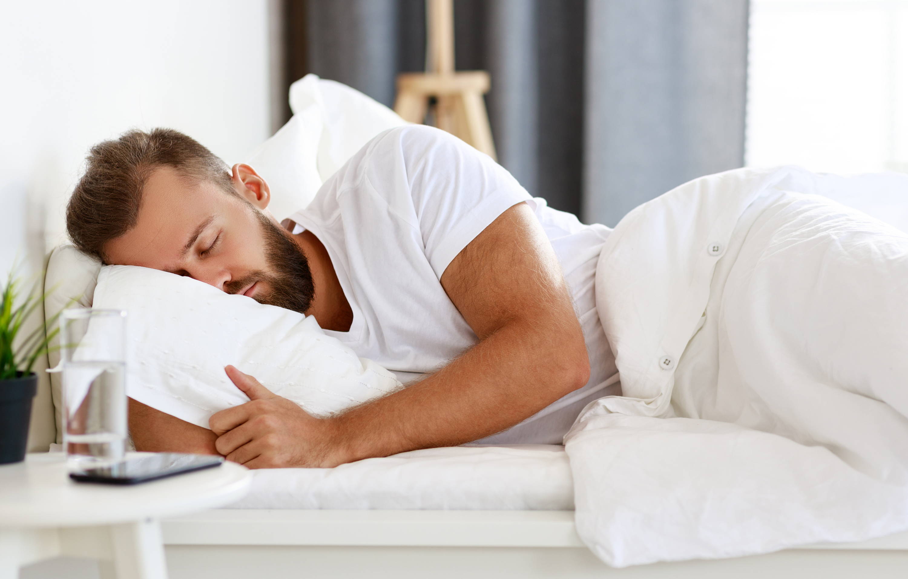 A man sleeping and cuddling with a white pillow and white blankets. 