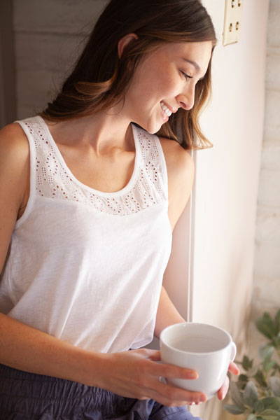 Woman leaning against door frame with coffee cup in hand wearing Seychelle tank top in white.