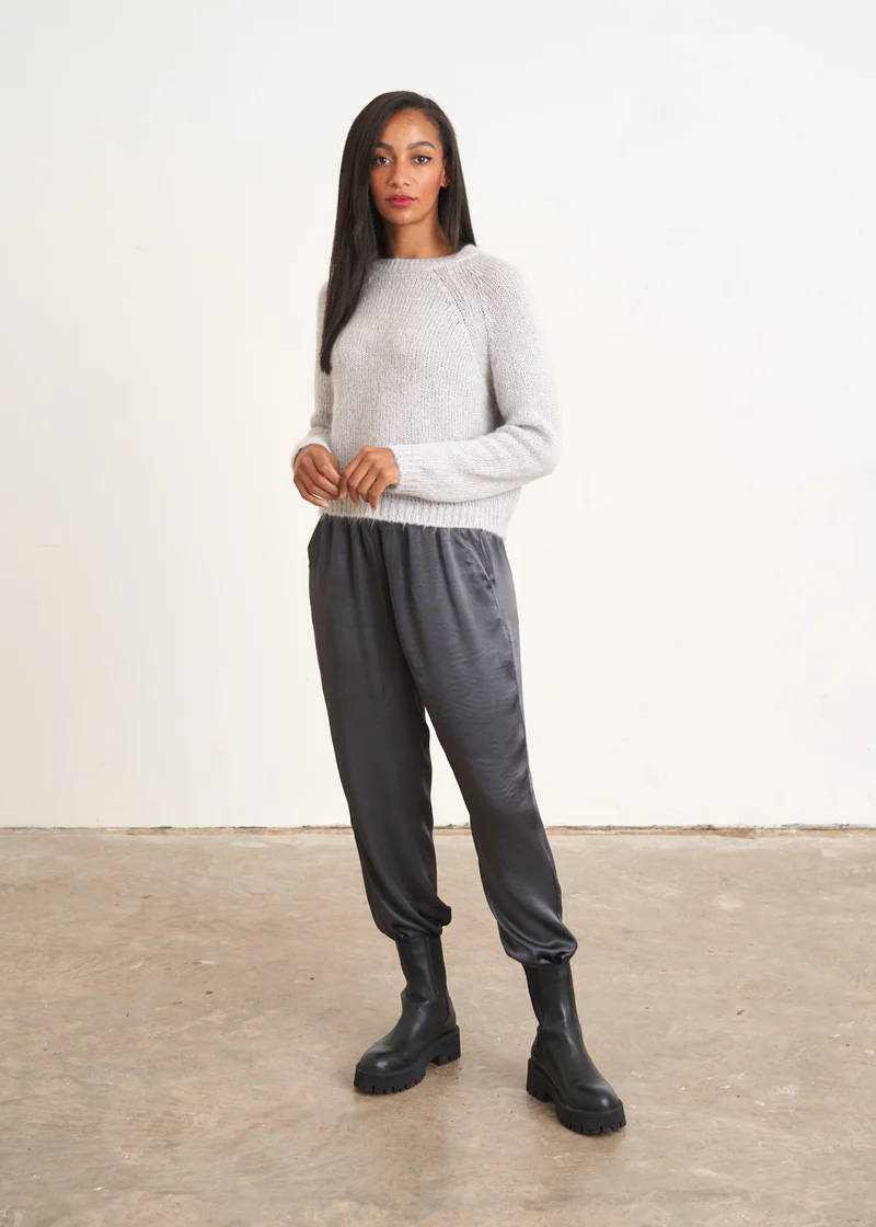 A model wearing a light grey knitted jumper over a pair of dark grey satin trousers and black leather chelsea boots