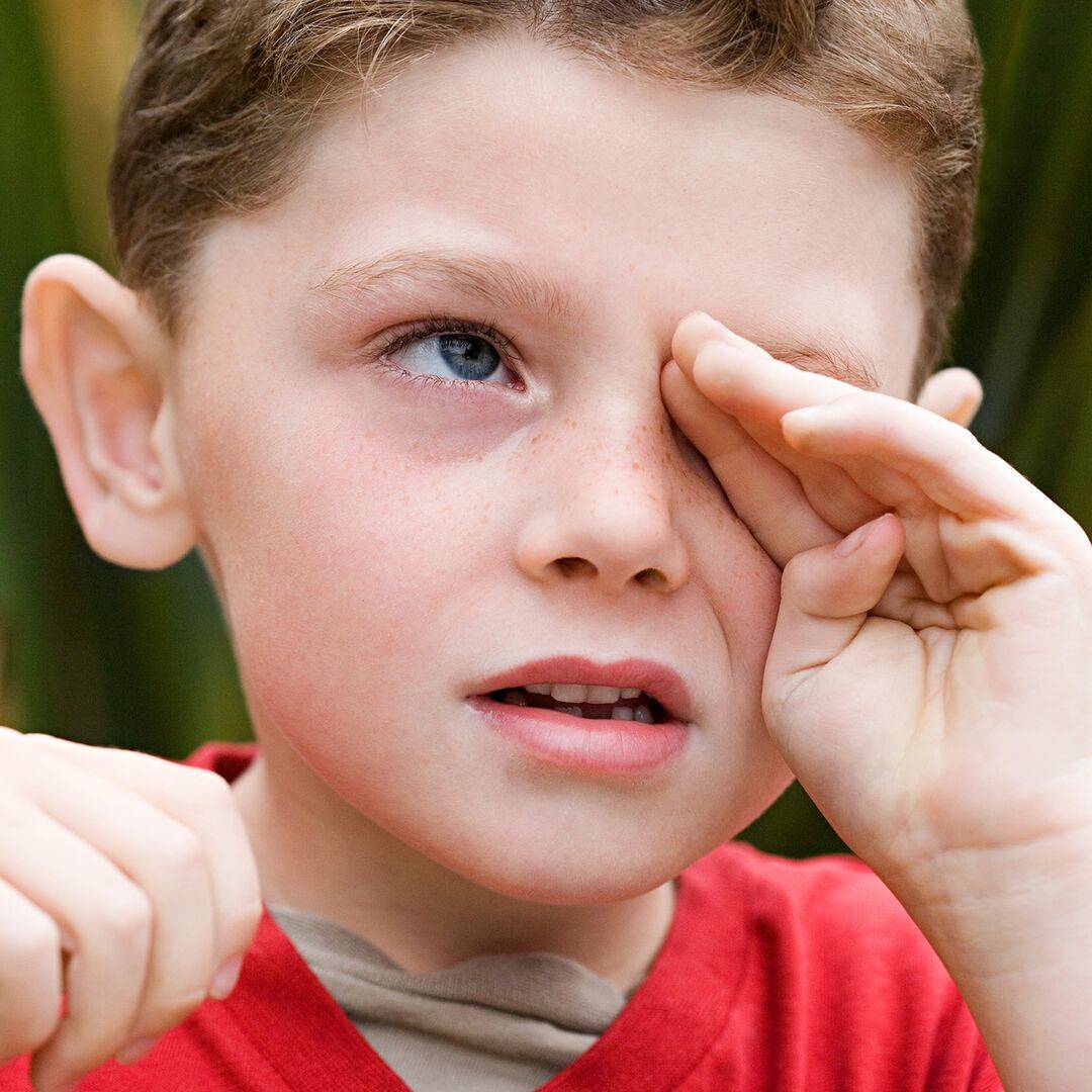 Ein Junge reibt sich das linke Auge mit der Hand - Juckreiz, Brennen und gerötete Augen sind häufige Allergiesymptome an den Augen bei Kindern.