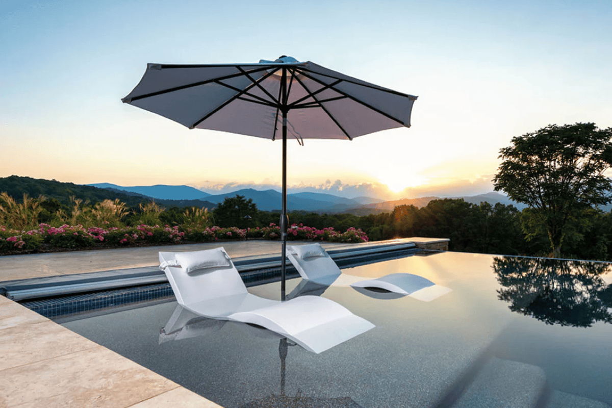 White in-pool chaise lounges and a patio umbrella on a sun shelf, designed for shaded poolside lounging and summer relaxation.