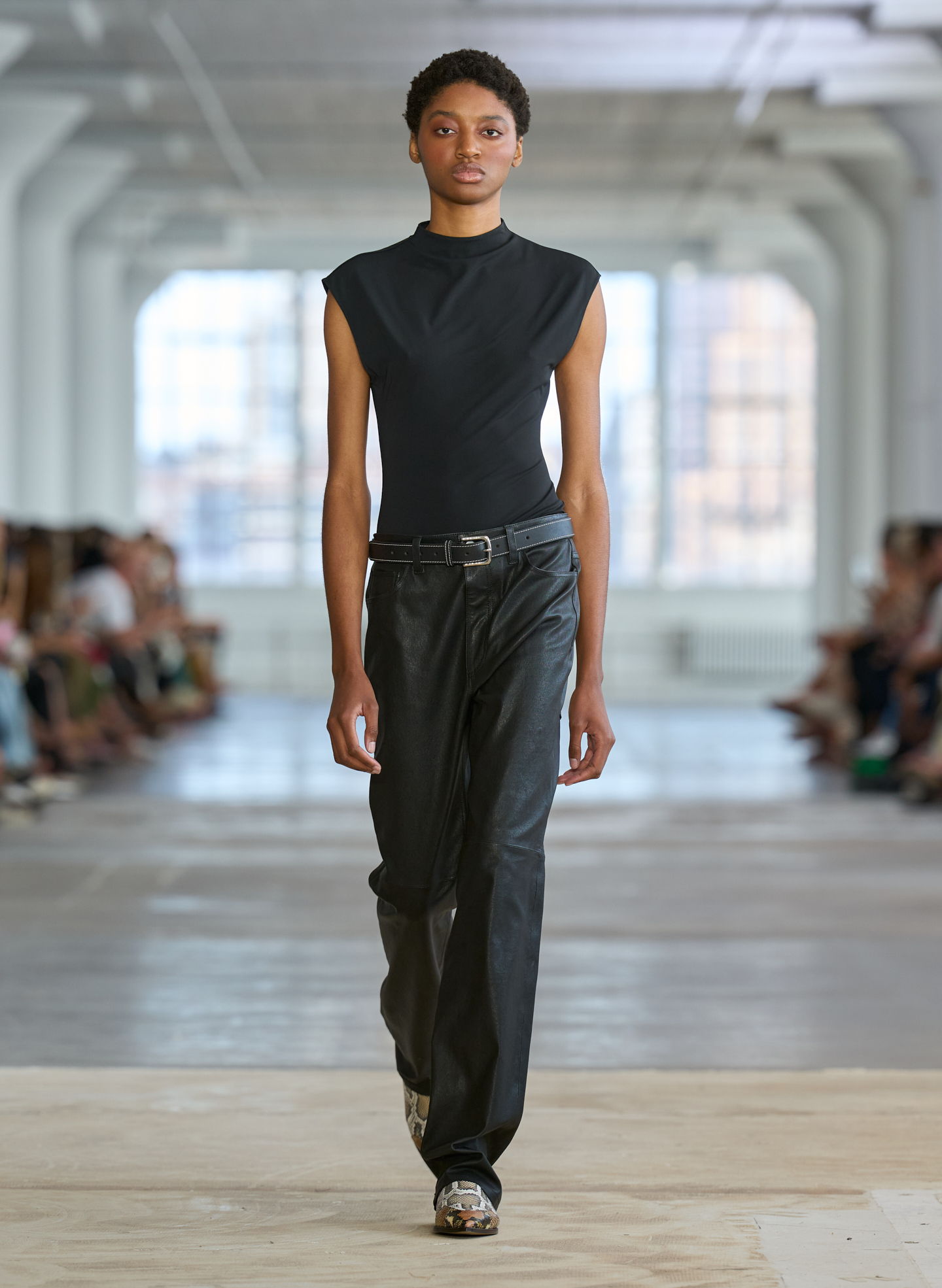 Model walking on runway wearing black outfit amidst seated audience.