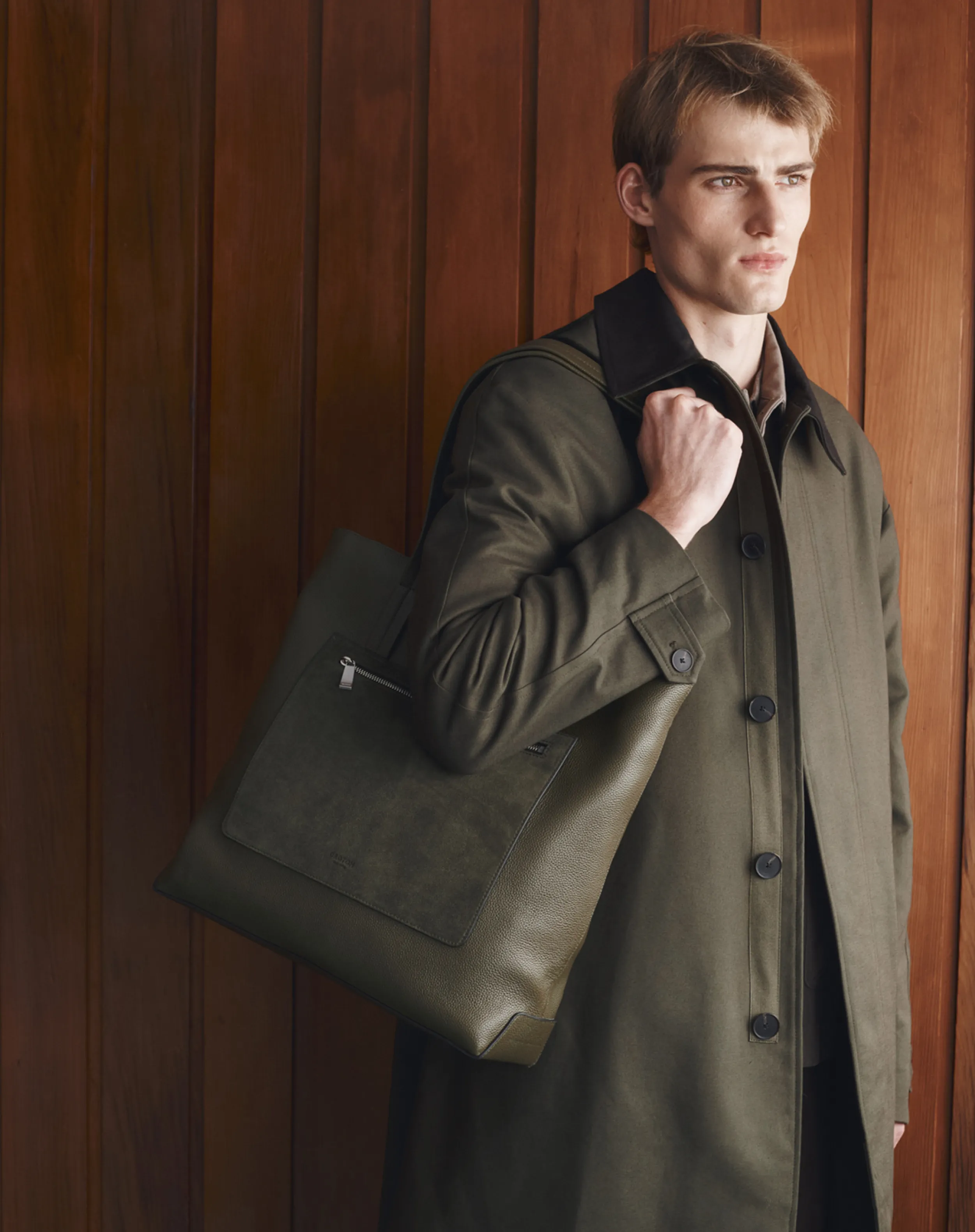 A man in a coat holding a leather bag against a wooden background.