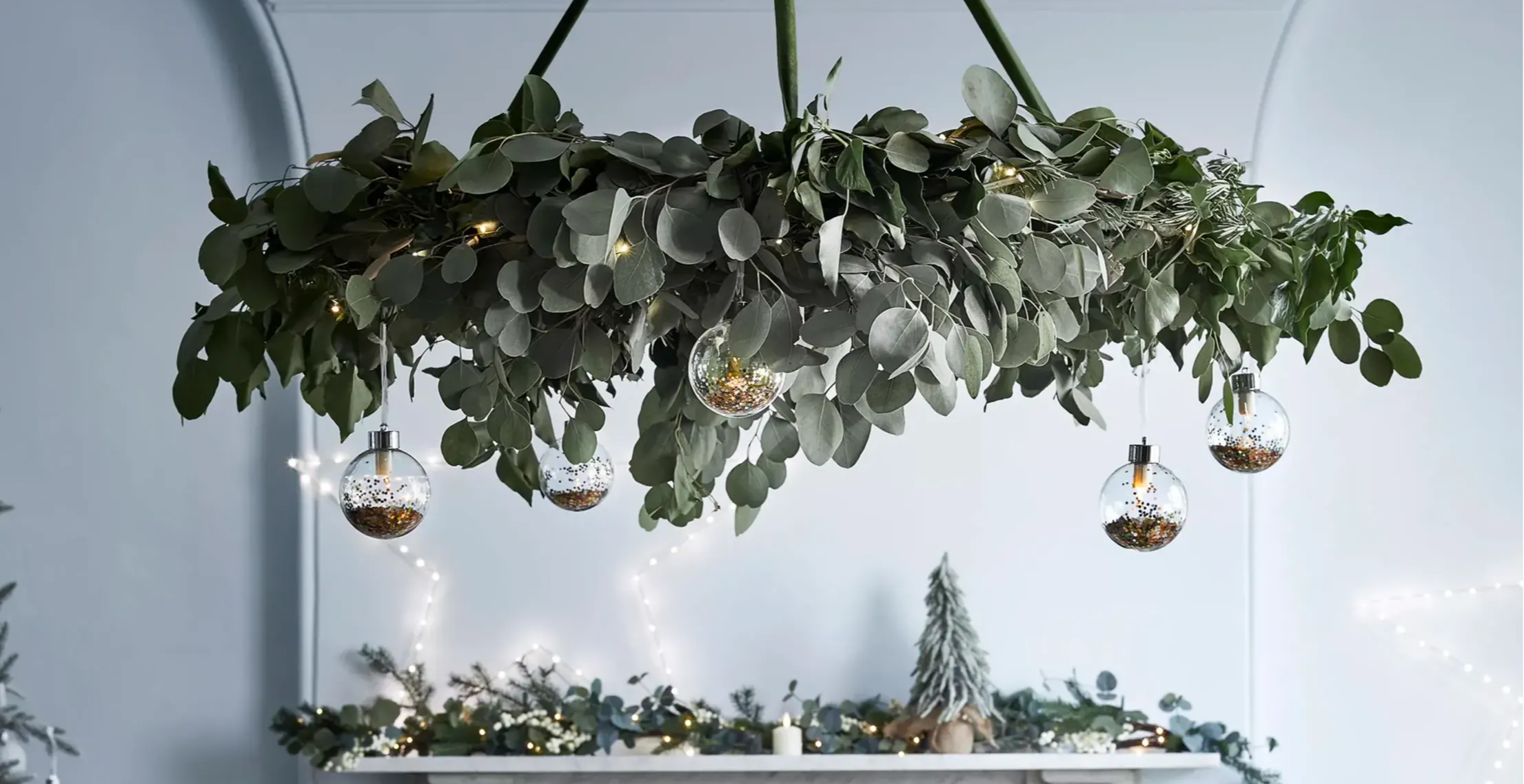 Close up image of foliage chandelier feature with mantel in the background featuring a festive garland, star light and mini tree