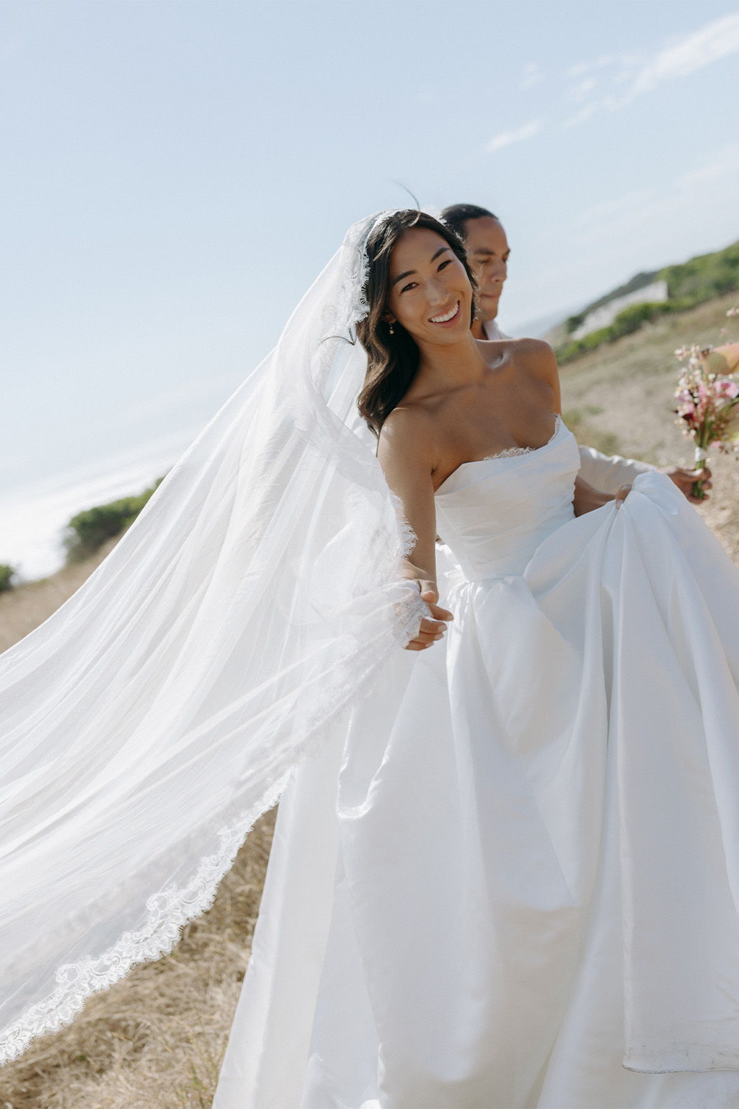 Bride in a white gown smiling with flowing veil in a scenic outdoor setting.