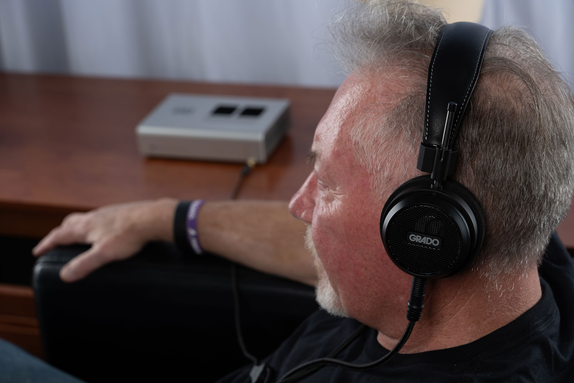 Photo taken from the side of a man relaxing in a black chair wearing the Grado S150 in front of a wooden table.