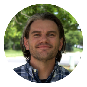 Smiling man with long hair, wearing a plaid shirt, outdoors in daylight.