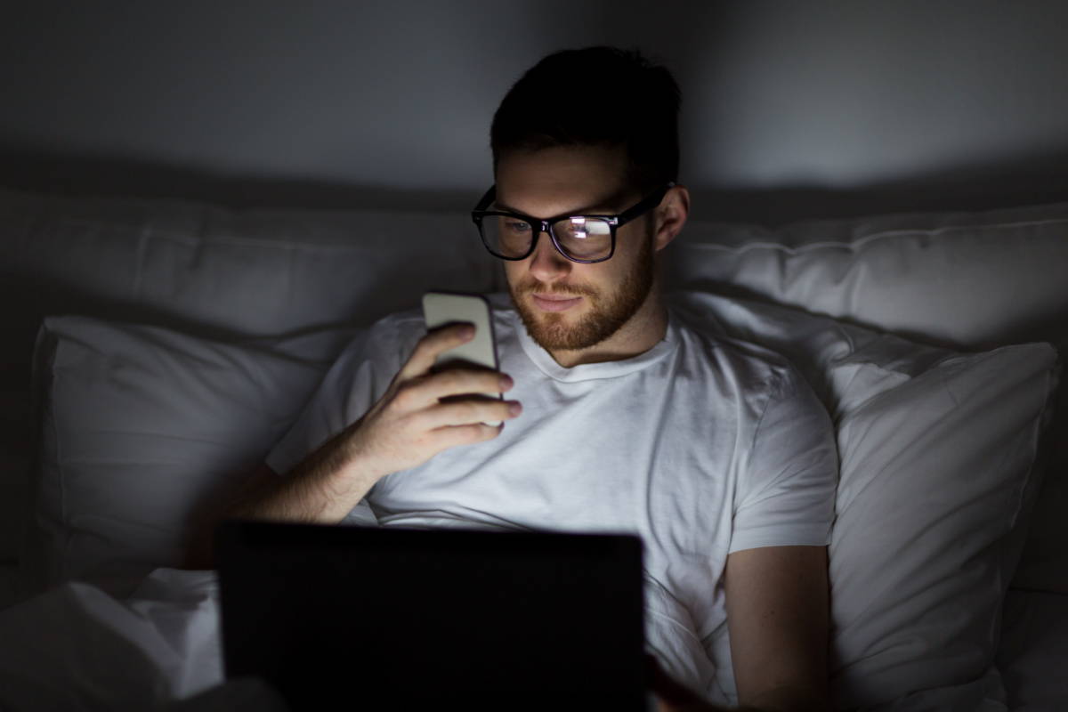 A man lounging in bed at night with a laptop on his lap and a phone in his hand 
