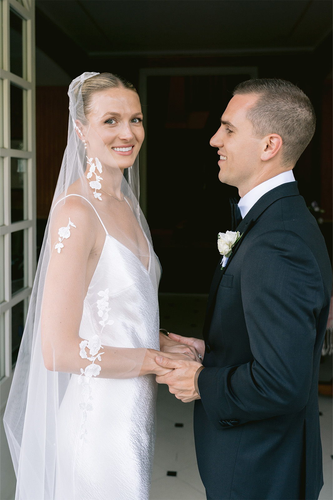 Bride and groom holding hands, smiling at each other, in a romantic setting.