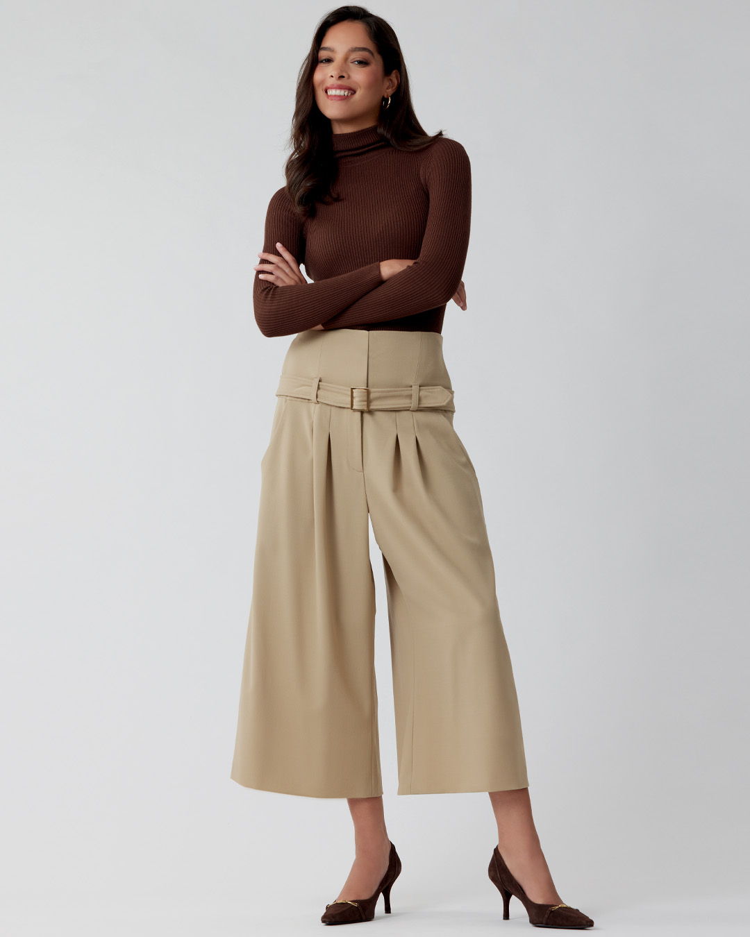 Woman in brown top and beige culottes, standing confidently with arms crossed.