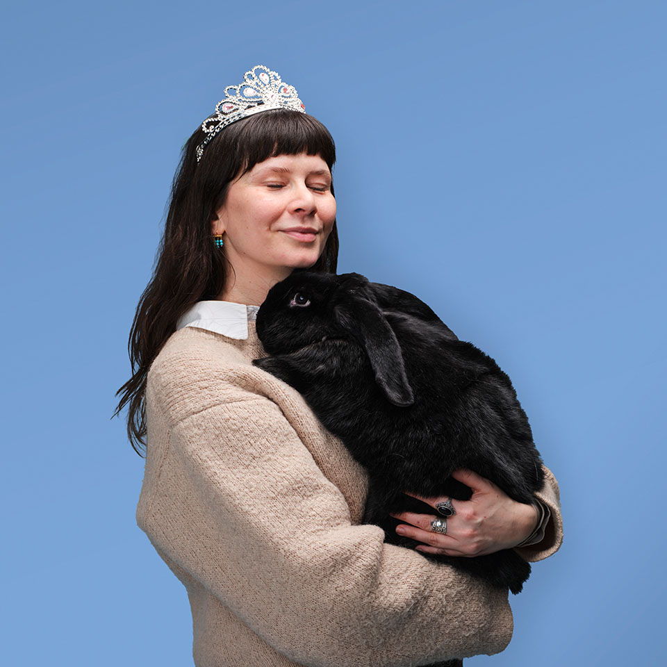Eine Person mit Krone, die ein großes schwarzes Kaninchen hält, vor blauem Hintergrund.