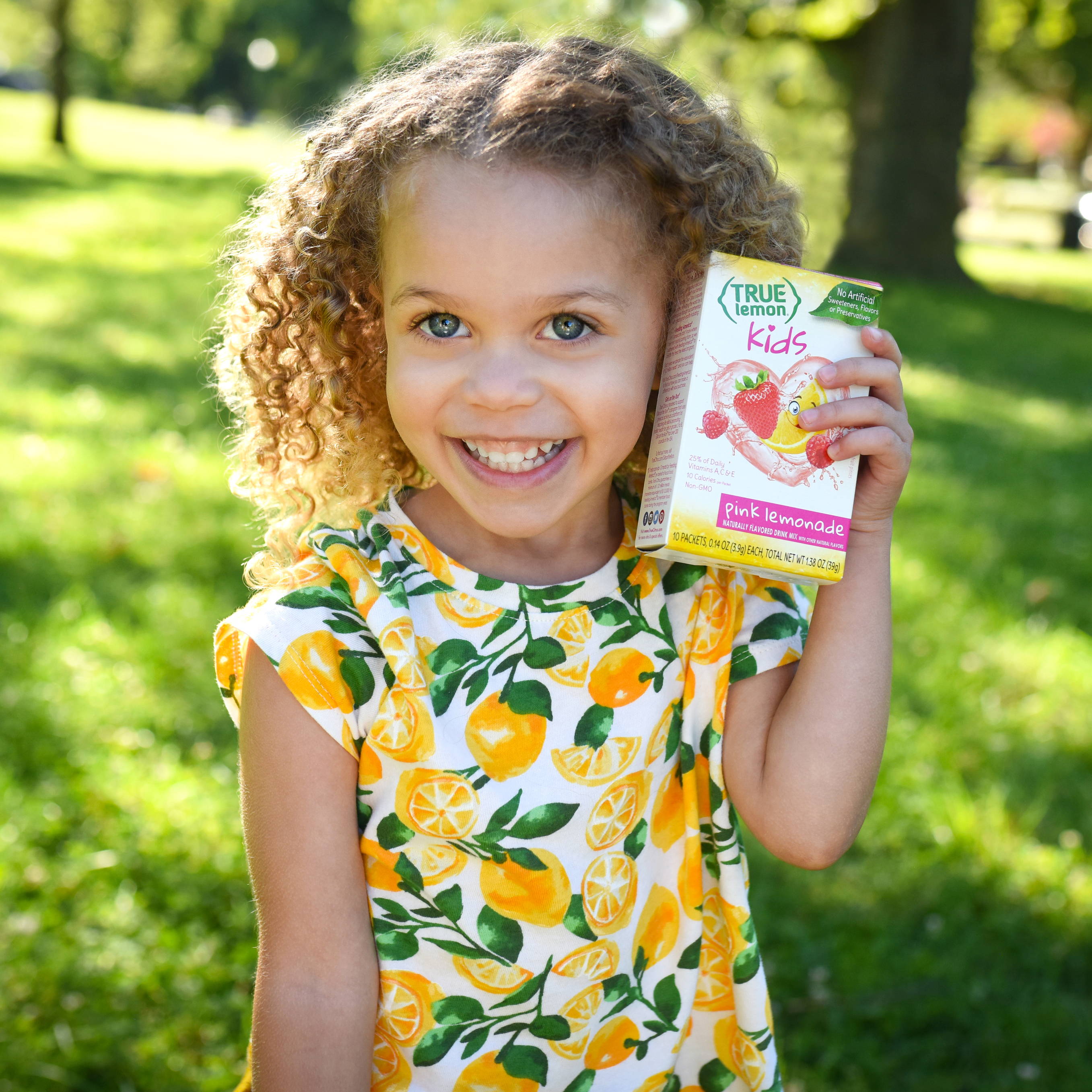 Girl in lemon print dress smiling while holding a box of true lemon kids strawberry lemon packets.
