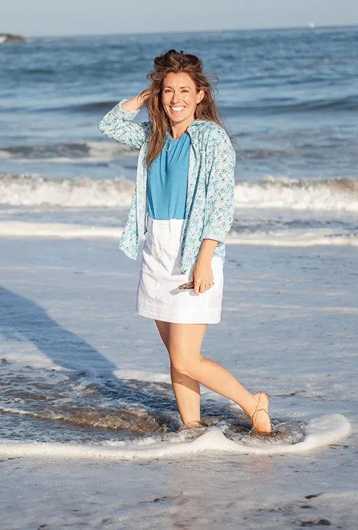 Smiling woman standing in shallow water at the beach, wearing casual clothes.