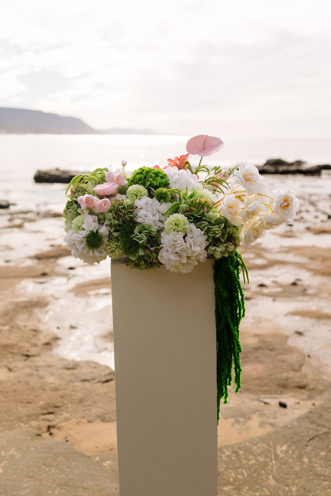 A floral arrangement on a pedestal by the ocean.