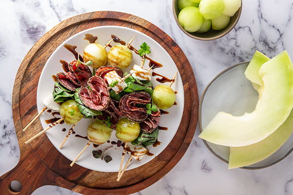 Skewers of meat and melon on a wooden board with additional melon slices.