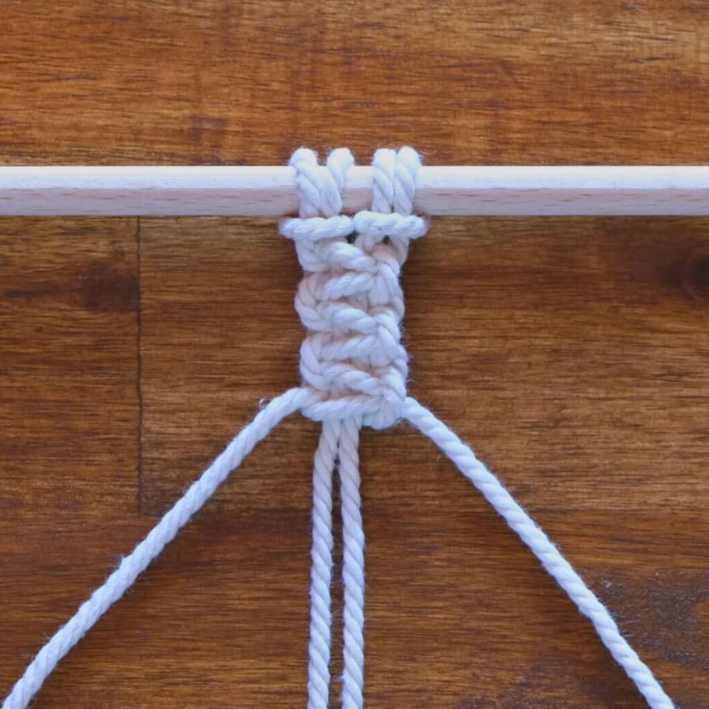 Ein Kreuzknoten, der mit einem weißen Seil um einen Holzstab auf einem Tisch gebunden ist.