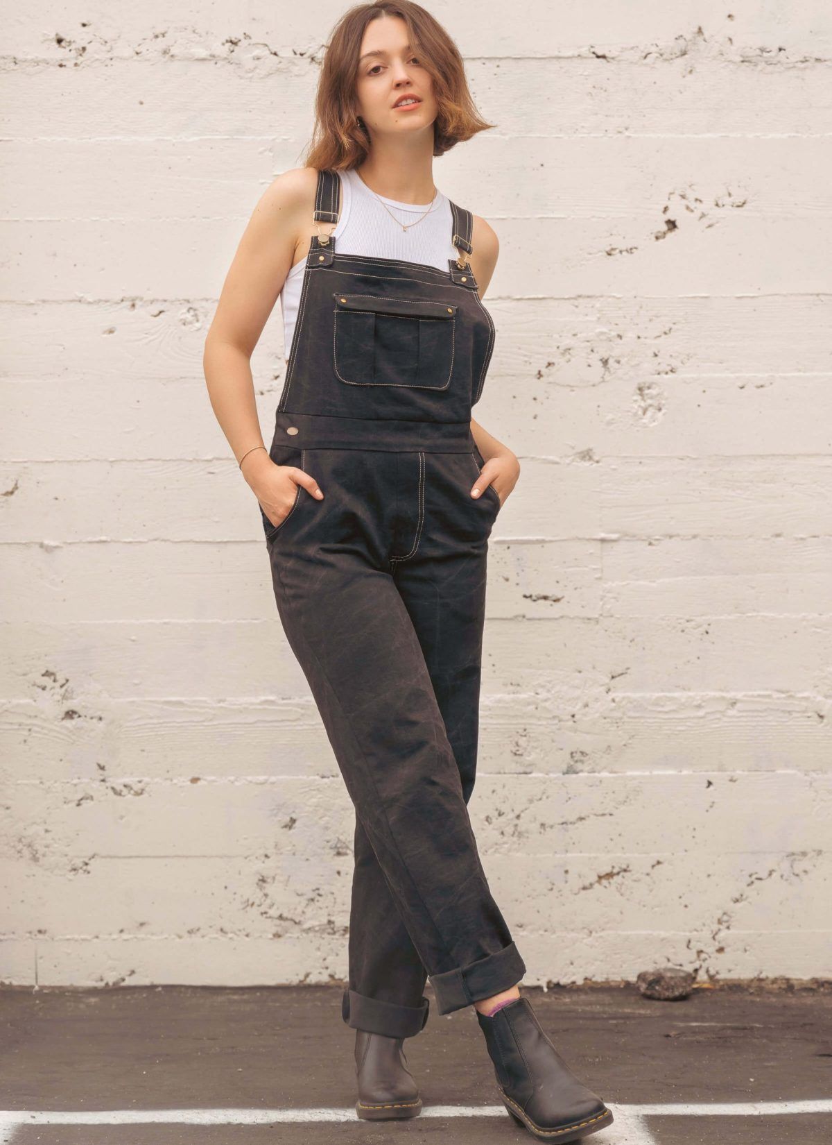 Person wearing black overalls and a white tank top, standing against a wall.