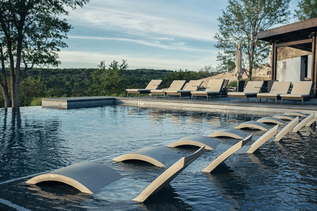 In-pool chaise lounges lined across a tanning ledge with poolside loungers in the background at a modern luxury outdoor space.