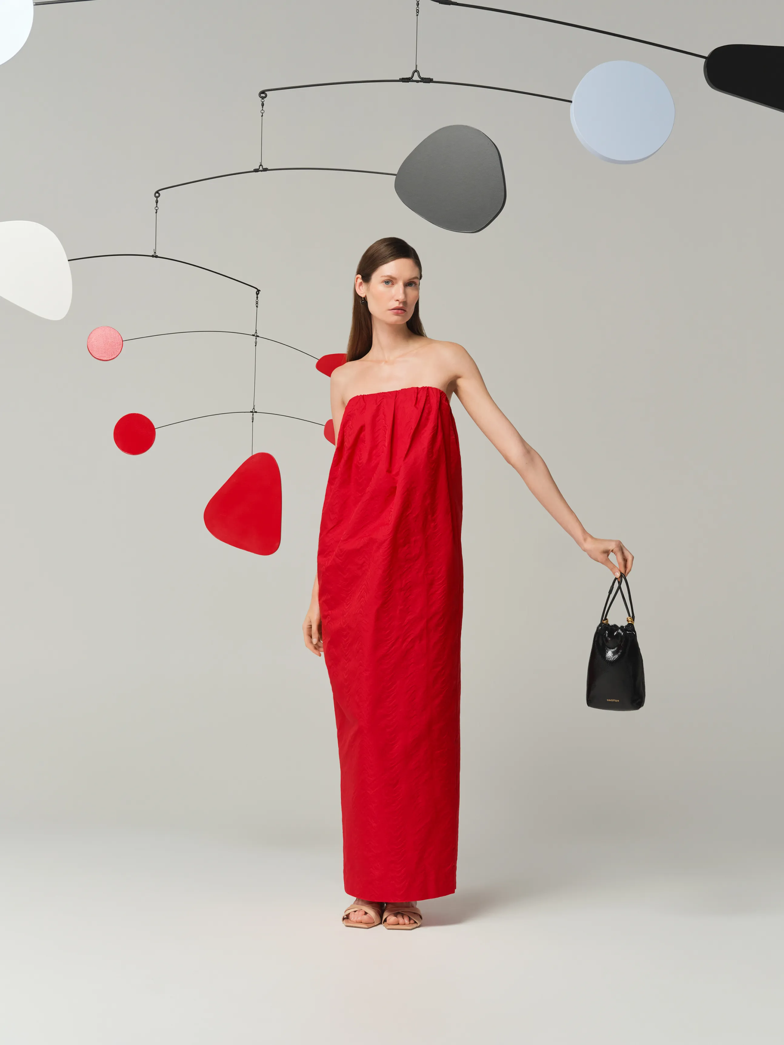 Model in a red dress stands with a black handbag against a minimalist backdrop.