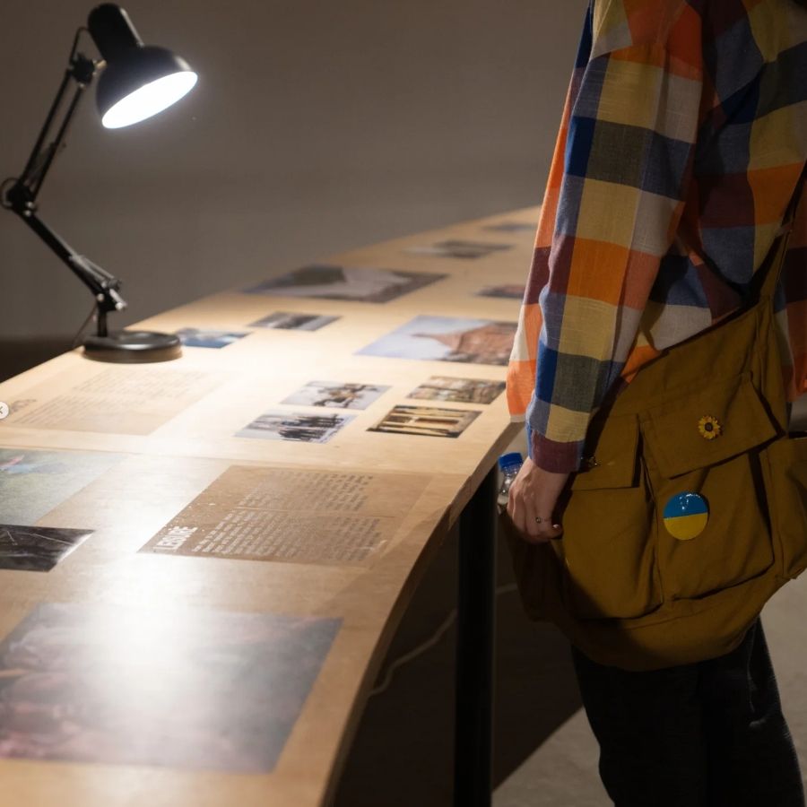 Person observing photographs on a table under a lamp in a gallery setting.
