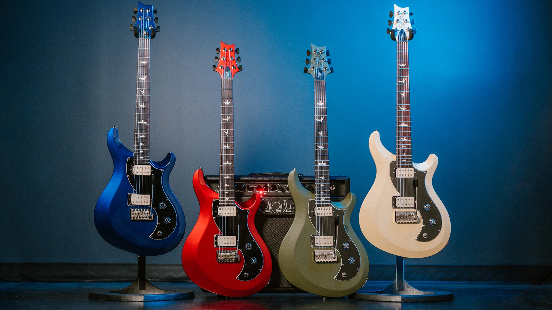 Four PRS S2 Vela HHT electric guitars in various colours displayed against a blue background.