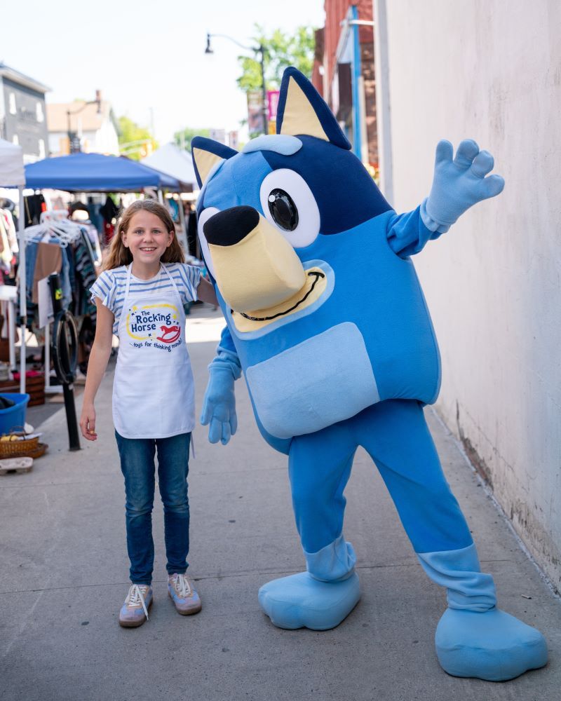 A girl poses with a blue cartoon character in a lively outdoor setting.