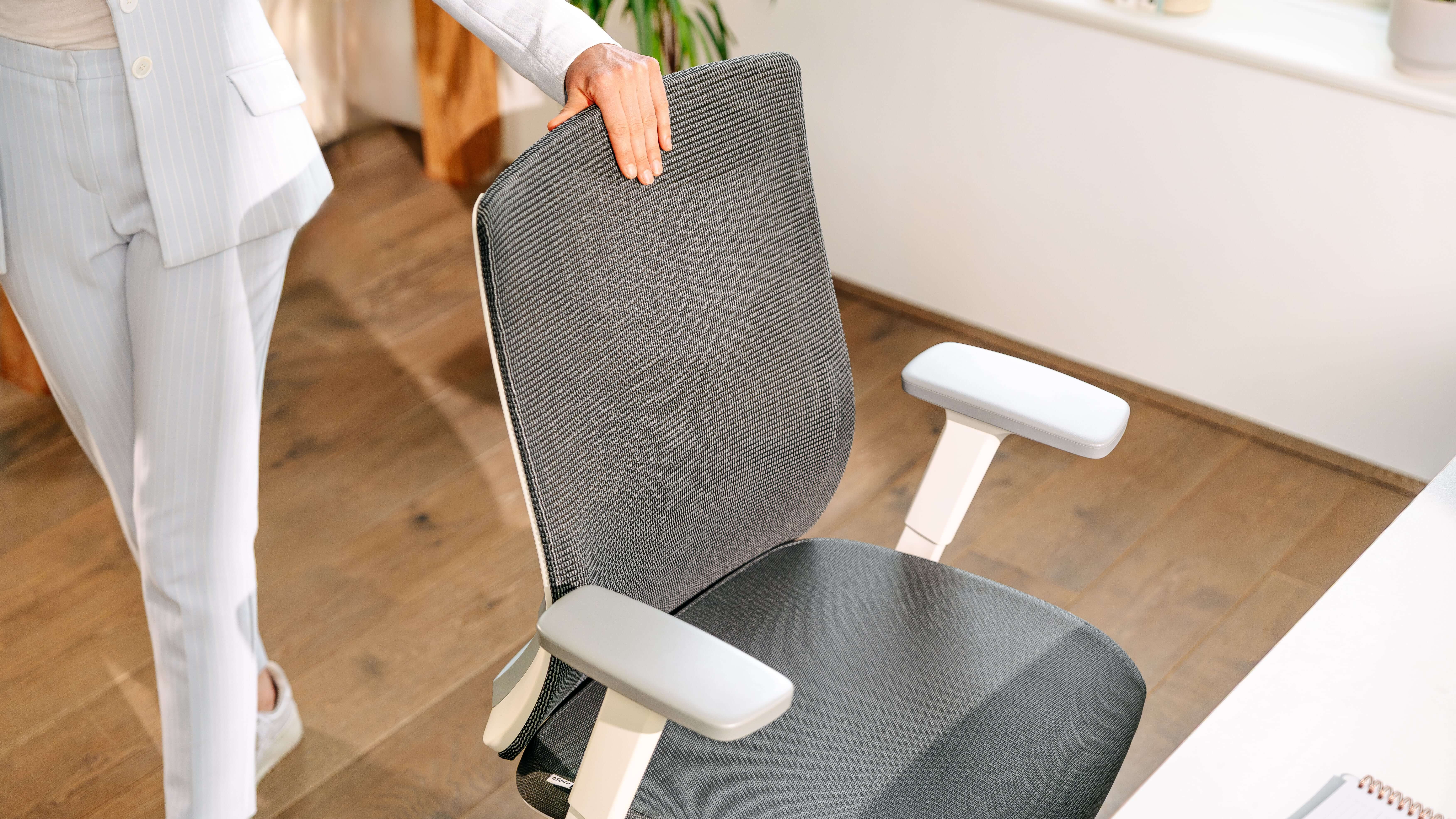 Une personne s'appuie contre le dossier en résille de la chaise de bureau ofinto Ergo dans un bureau à domicile lumineux doté d'un parquet.