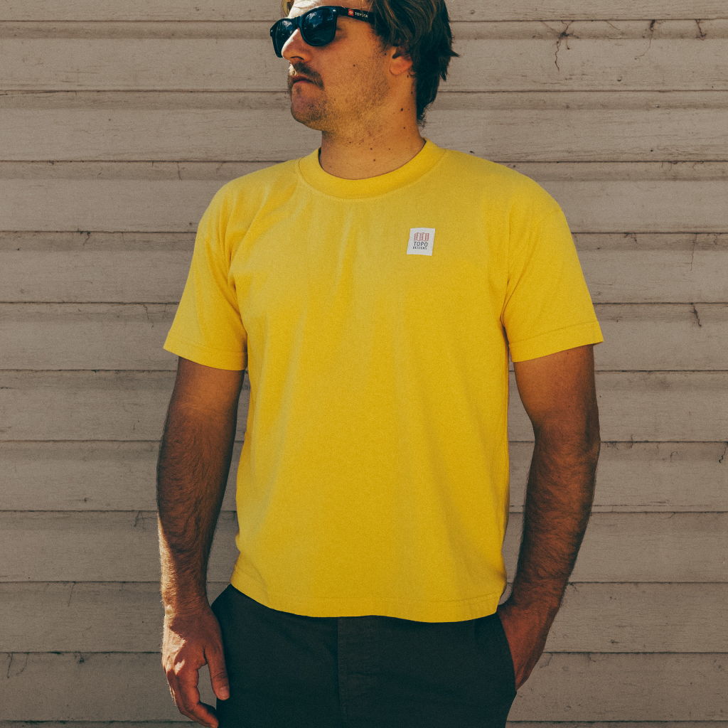 Man wearing a yellow t-shirt and sunglasses, standing against a wooden background.