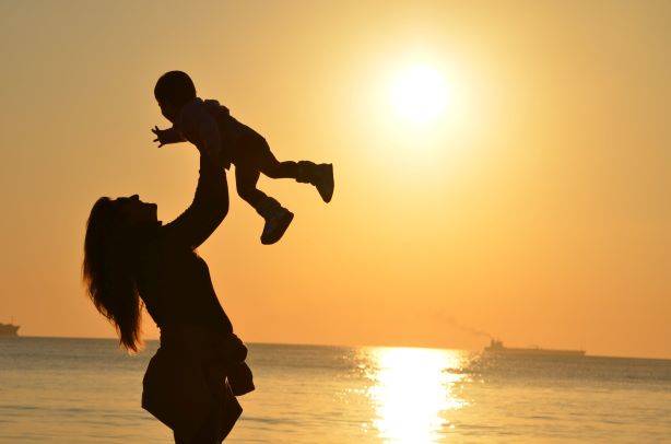 Woman Holding Baby Up In The Air By The Sea & Sunset