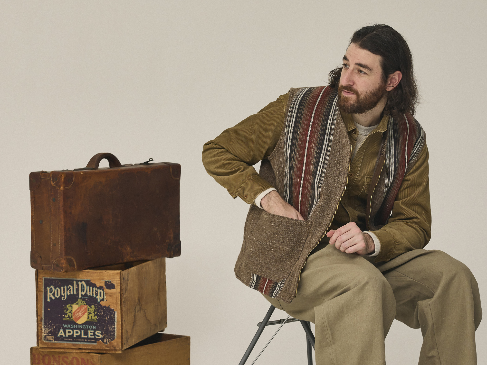 Man sitting on chair next to stacked wooden suitcases against plain background.