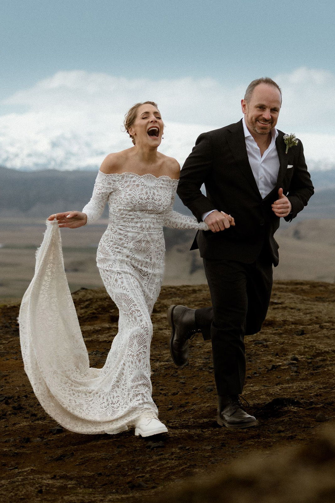 Bride and groom joyfully running outdoors, wearing wedding attire, with mountains in background.