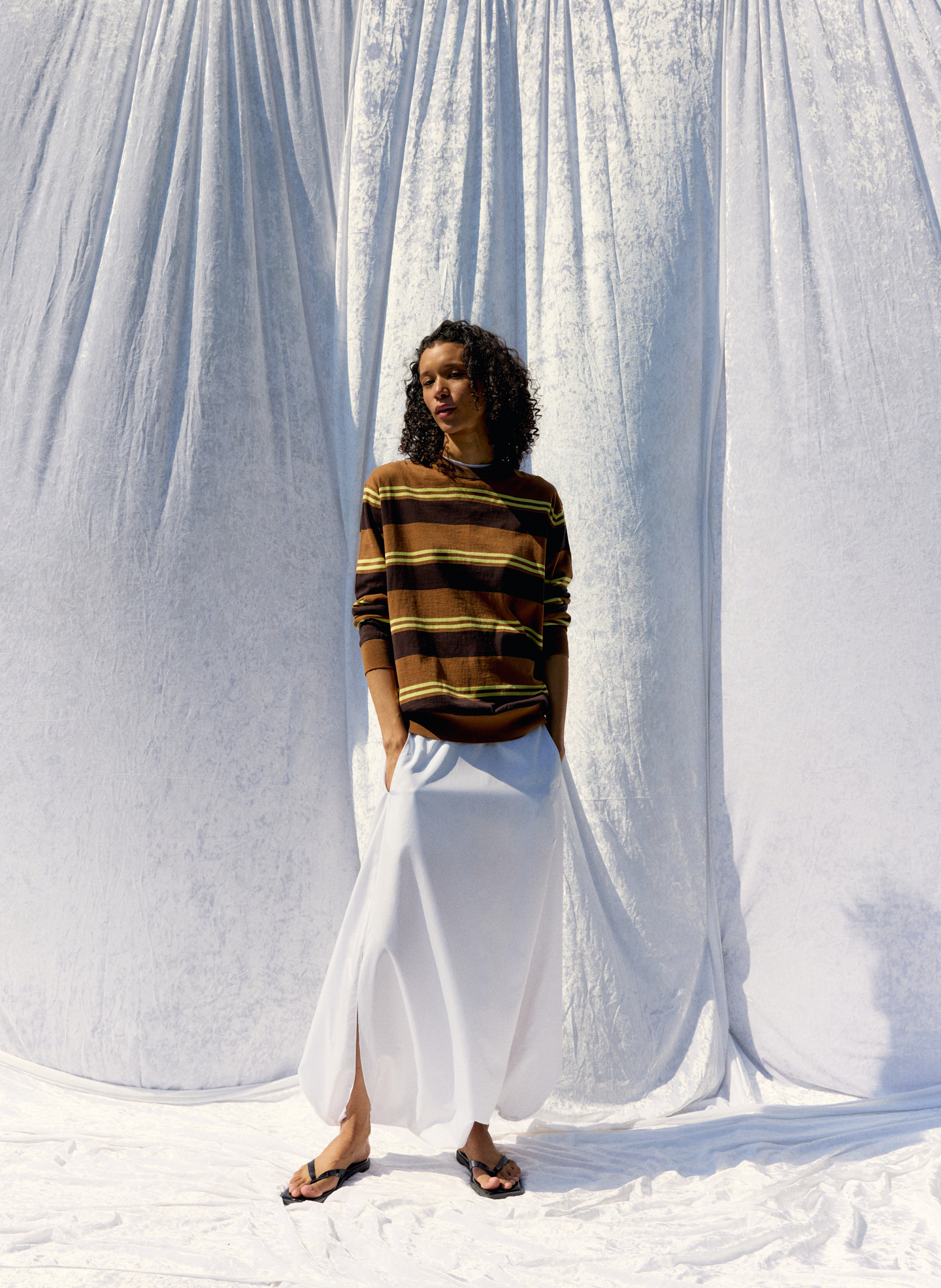 Person in striped sweater and white skirt standing against a draped background.