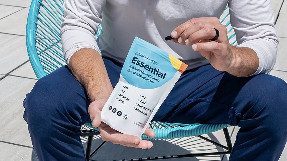 Plant-based founder Matt Tullman sitting in chair holding Complement Essential vegan multivitamin pouch and capsules. 