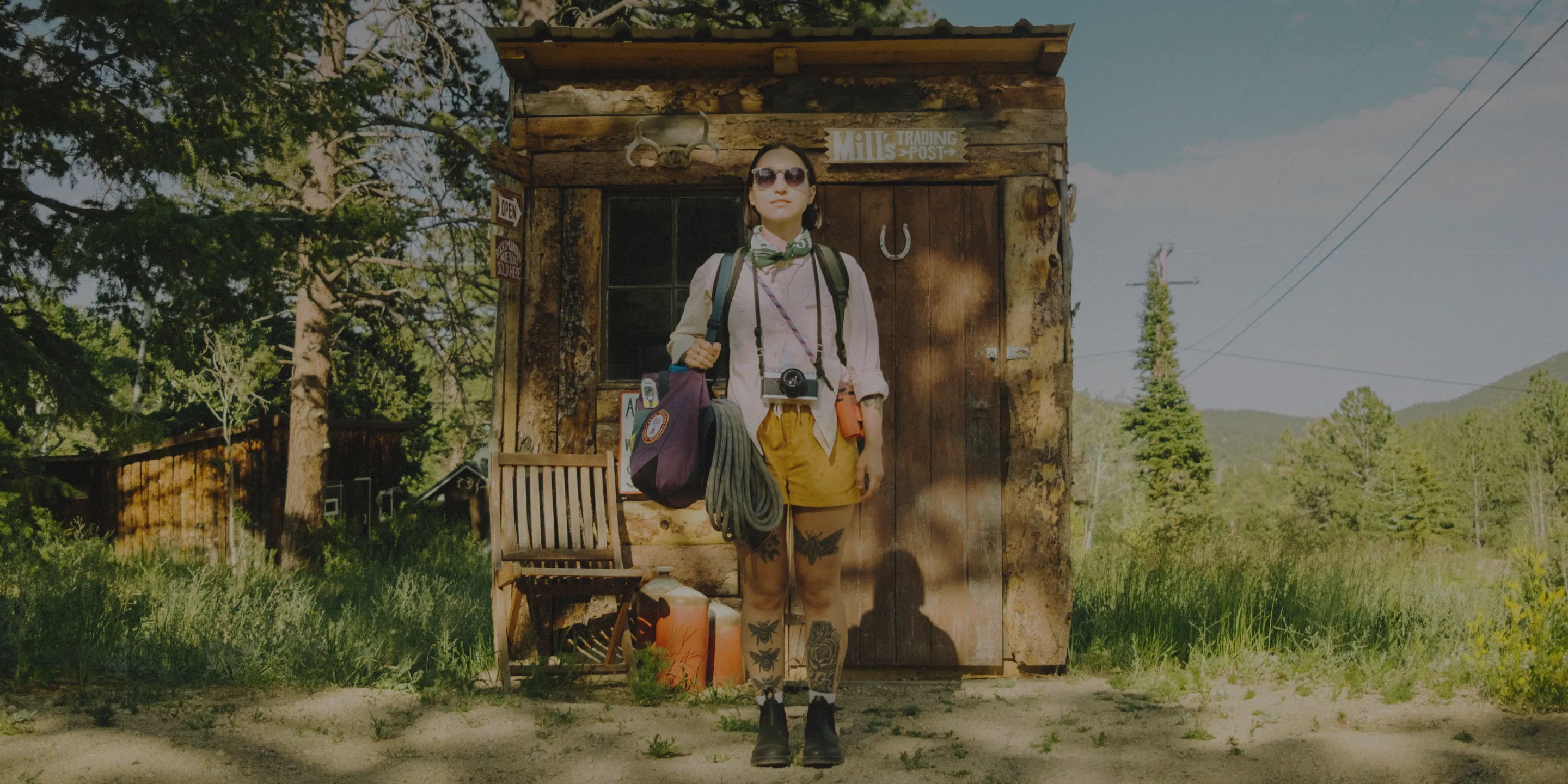 Person standing in front of a cabin, holding bags in a forested area.