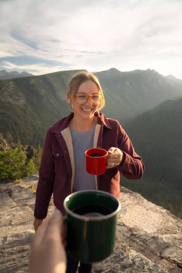 Two women cheers coffee cups on cliff top over looking valley with sunset.